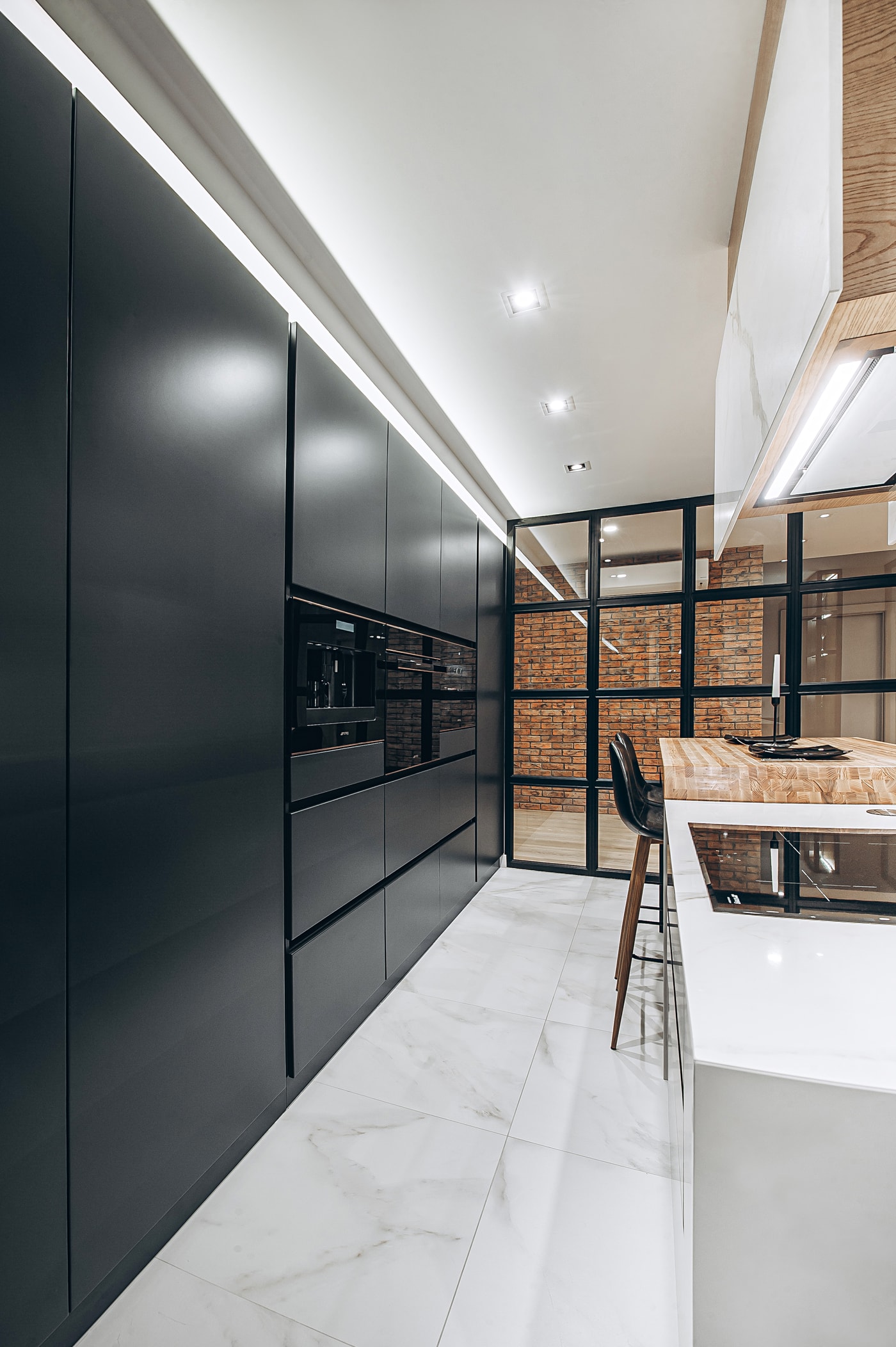 Full-height black matte cabinet fronts with an LED cove and built-in appliances, a brick wall behind the glass