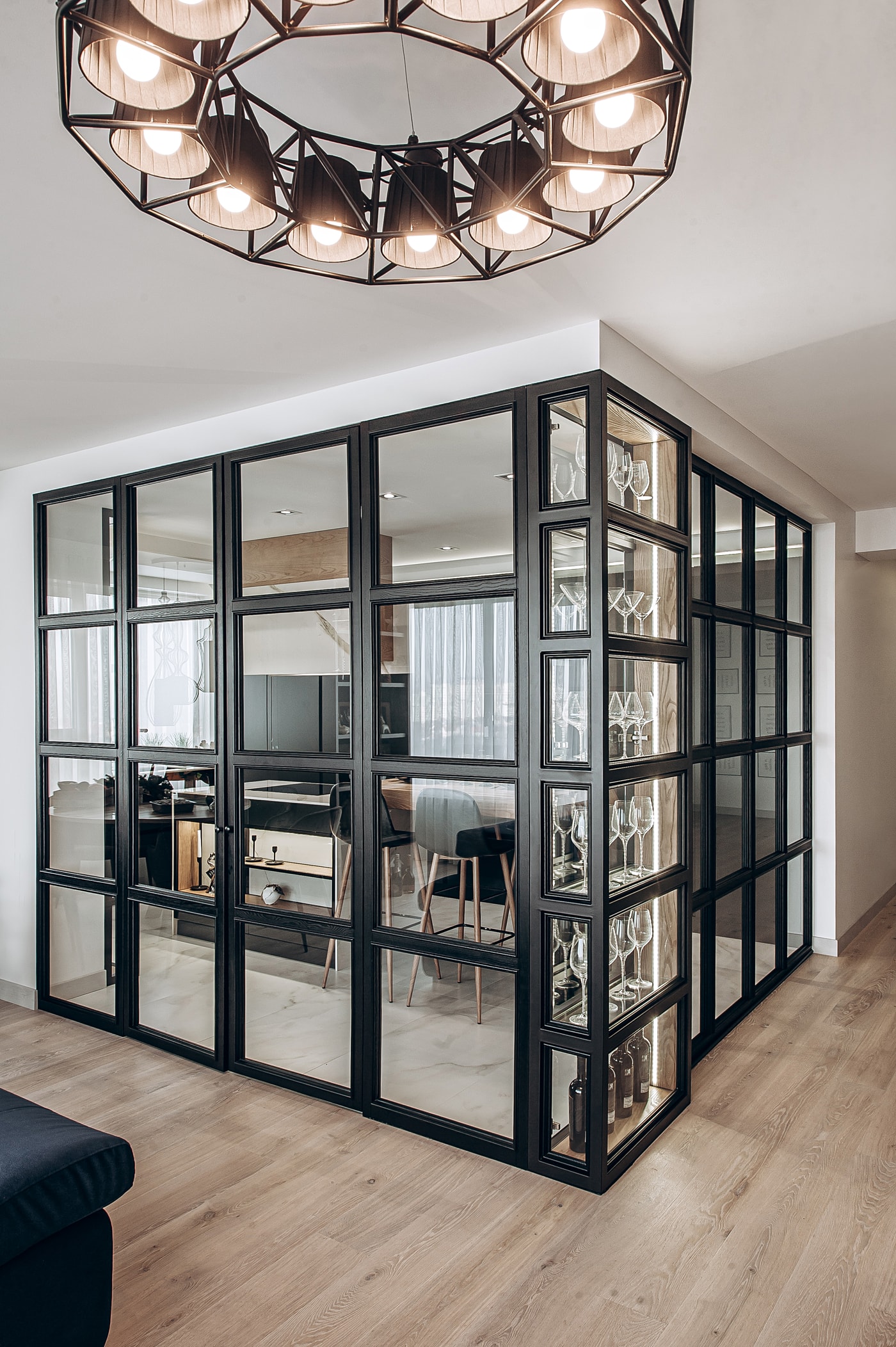Corner glass partition with a glassware display and a pendant chandelier, seen from the living room