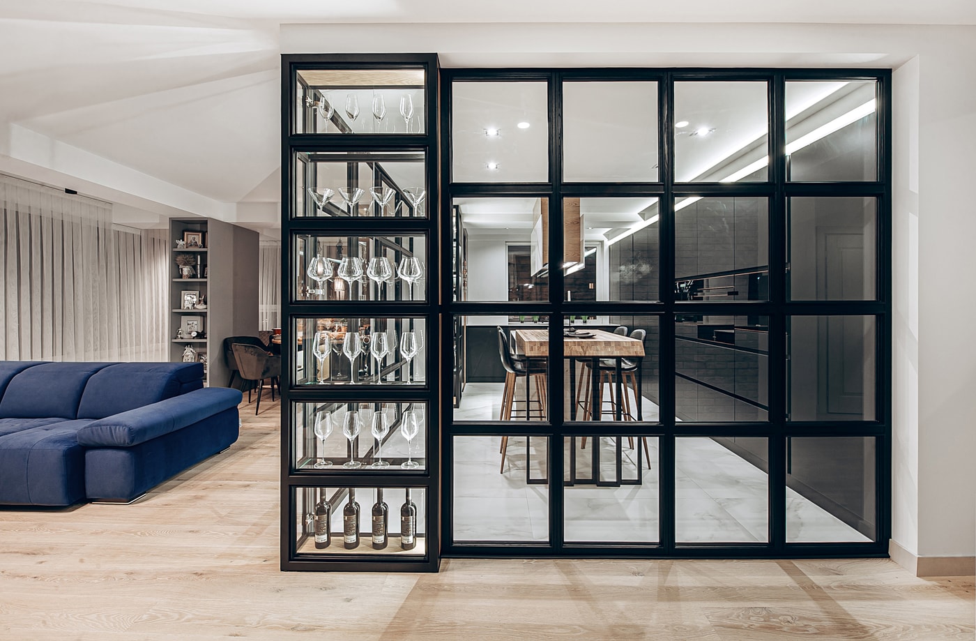 Glass partition in a black frame with an illuminated glassware display, looking from the living room into the kitchen